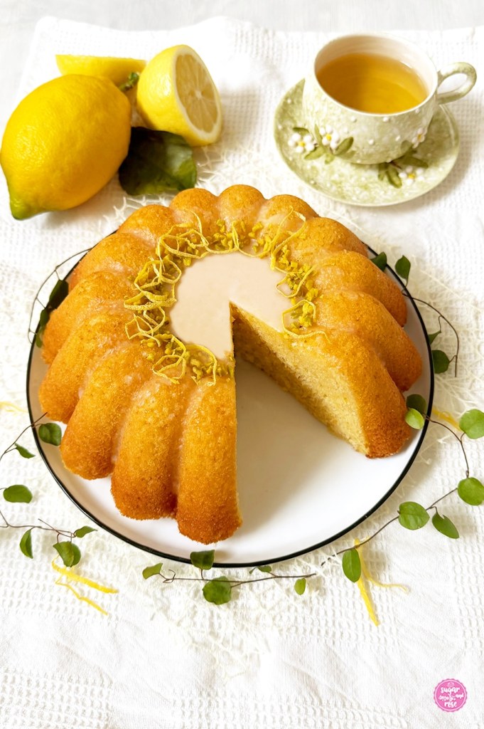 Getränkter Zitronengugelhupf angeschnitten, dahinter Zitronen mit grünem Zitronenblatt und eine Schale grüner Tee in hellgrüner Keramik mit weißen haptischen Blüten darauf