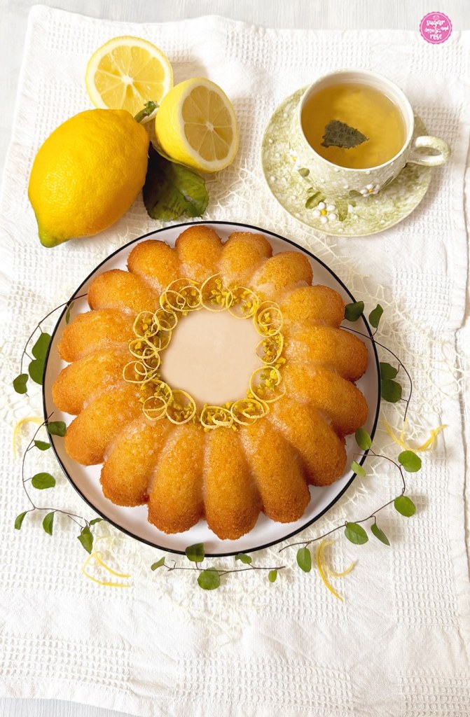Getränkter Zitronengugelhupf auf weißem Keramikteller mit zartem blauem Rand auf weißem geripptem Geschirrtuch, im Hintergrund eine ganze Zitrone mit grünem Blatt und zwei Zitronenhälften, daneben eine Schale grüner Tee