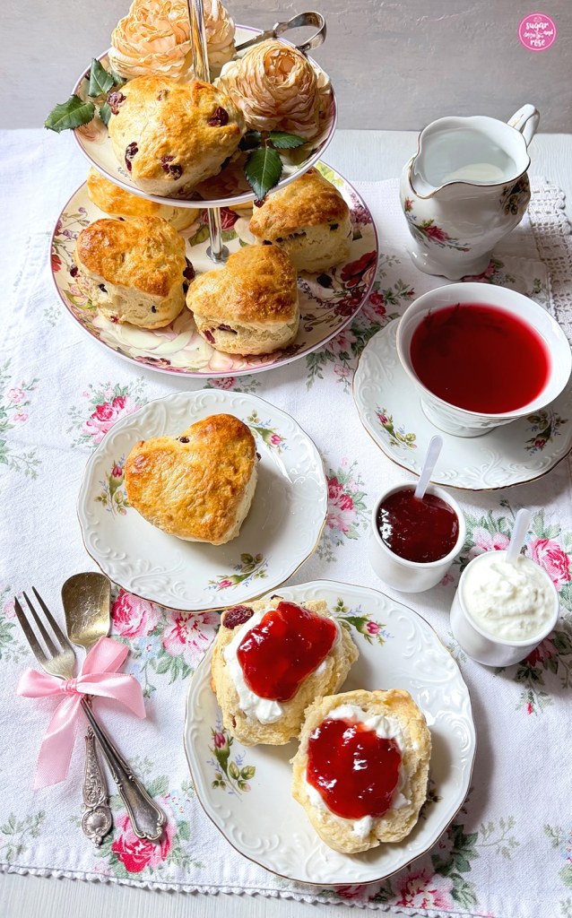 Cranberry-Scones auf Etagere mit zwei cremefarbenen Rosenblüten, eine Tasse roter Tee, ein Milchkännchen und im Vordergrund ein halbierter Scone mit Double Cream und Marmelade