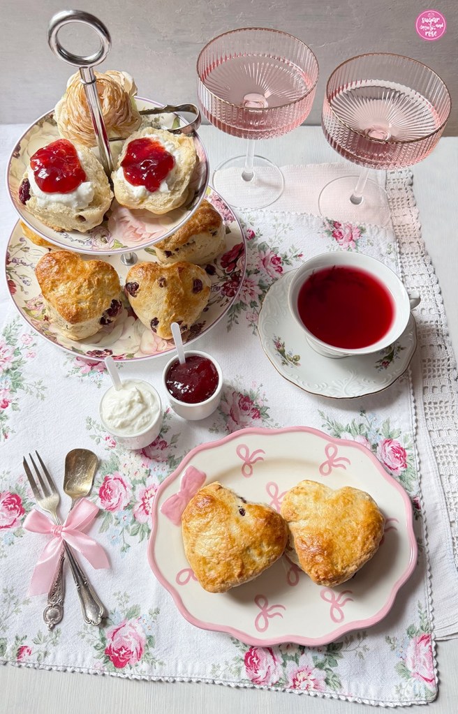 Scones in Herzform auf Etagere, davor ein ovaler Teller mit rosa Maschen als Dekor, darauf zwei herzförmige Cranberry-Scones, dazu eine Tasse Tee, zwei Gläser Lillet Berry und Erdbeermarmelade und Double Cream