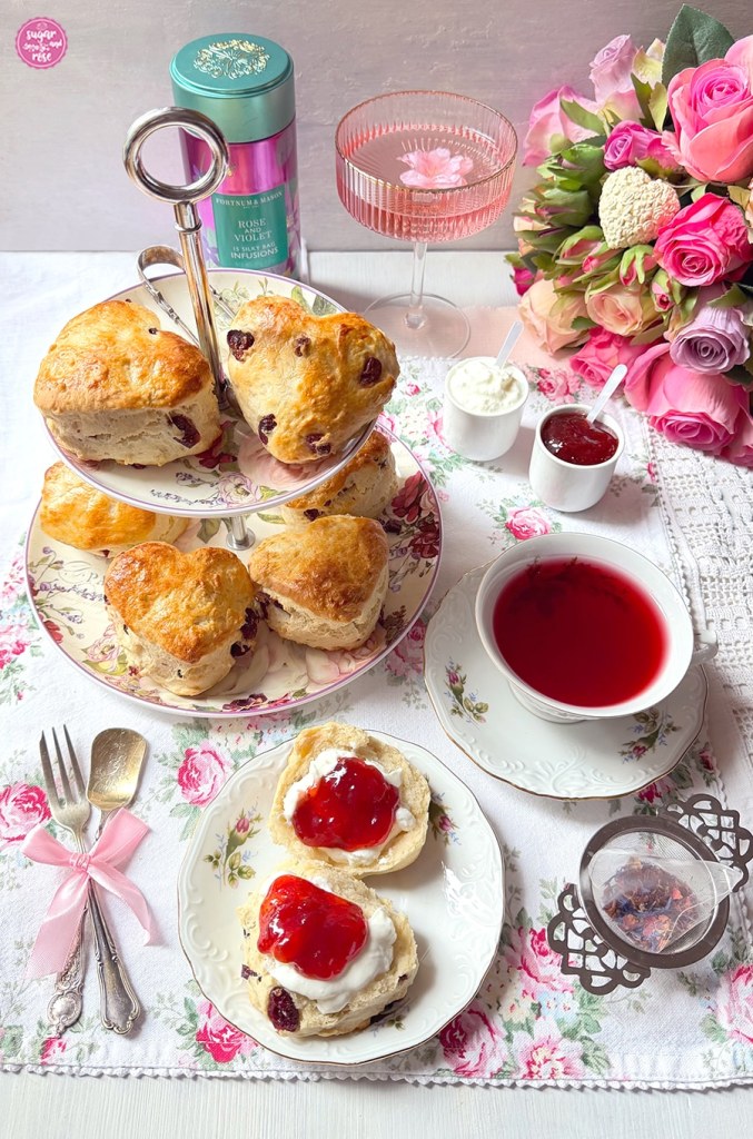 Cranberry-Scones auf zweistufiger Etagere mit Rosendekor, davor ein Dessertteller mit Rosenmuster und einem aufgebrochenen Scone mit Erdbeeermarmelade und Double Cream. Im Hintergrund ein Rosenstrauß in Rosatönen, eine Schale Lillet Rose und eine Teedose von Fortnum & Mason