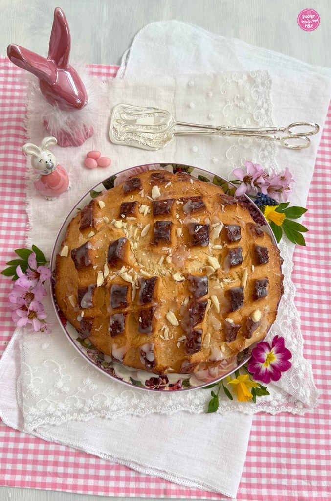Eine Osterflade ohne Rosinen, dafür mit Mandelstücken bestreut auf runden Teller mit Rosendekor, rundum rosa Blüten von Steinbrech, Primeln und kleinen gelben Narzissen, zwei rosa Prozellanhasen, einer mit weißem Pelzchen um den Hals und eine silberne Kuchenzange