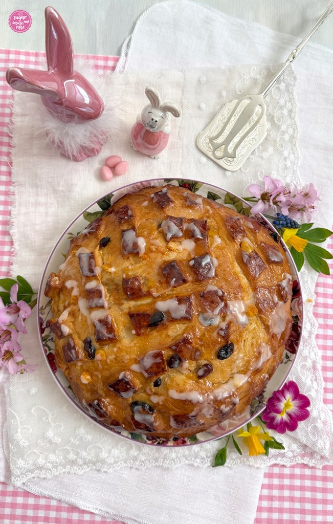 Osterfladen mit Zuckerglasur auf rosa-weiß karierter Stoffserviette, dazu eine silberene Kuchenzange und zwei rosa Porzeallanosterhasen