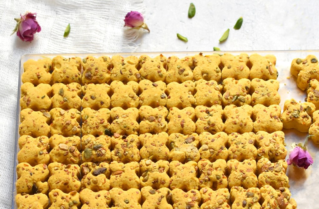 Kichererbsen-Kekse mit Pistazien dicht aneinandergereiht auf einem Glastablett, bestreut mit getrockneten rosa Rosenblüten und grünen Pistazienstiften