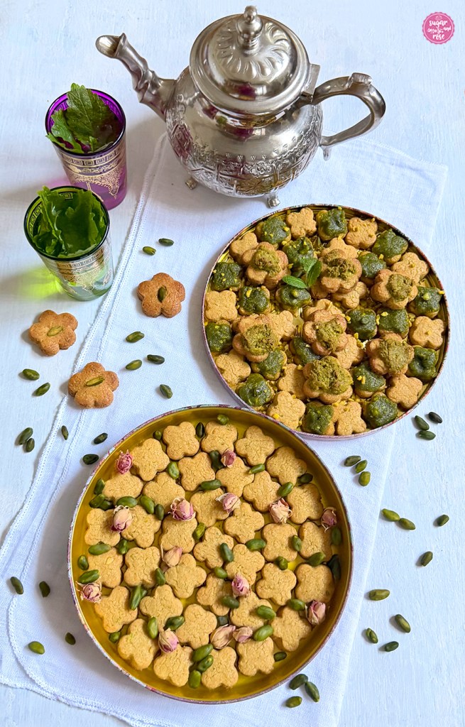 Kichererbsen-Kekse in zwei runden Blechschalen, einmal plan und einmal mit Pistazienpaste bestrichen, dazu eine silberne Teekanne und zwei orientalische Teegläser mit Pfefferminztee