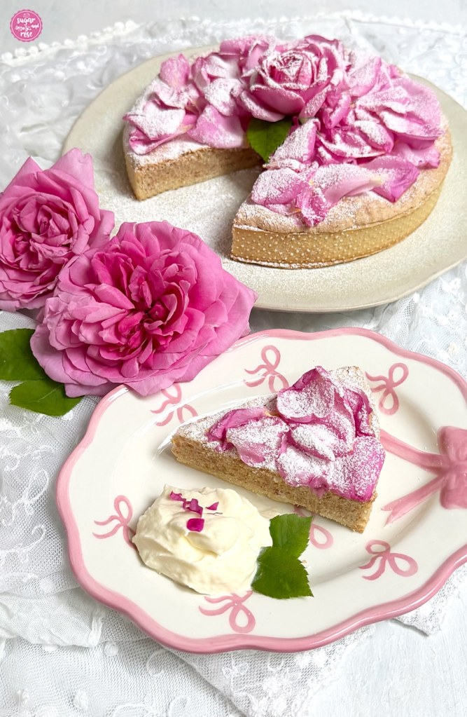 Ein Stück Mandelkuchen aus der runden Tarte herausgeschnitten auf einem ovalen Teller mit rosa Dekormaschen, dazu ein Klecks Sahne mit Rosenblüten