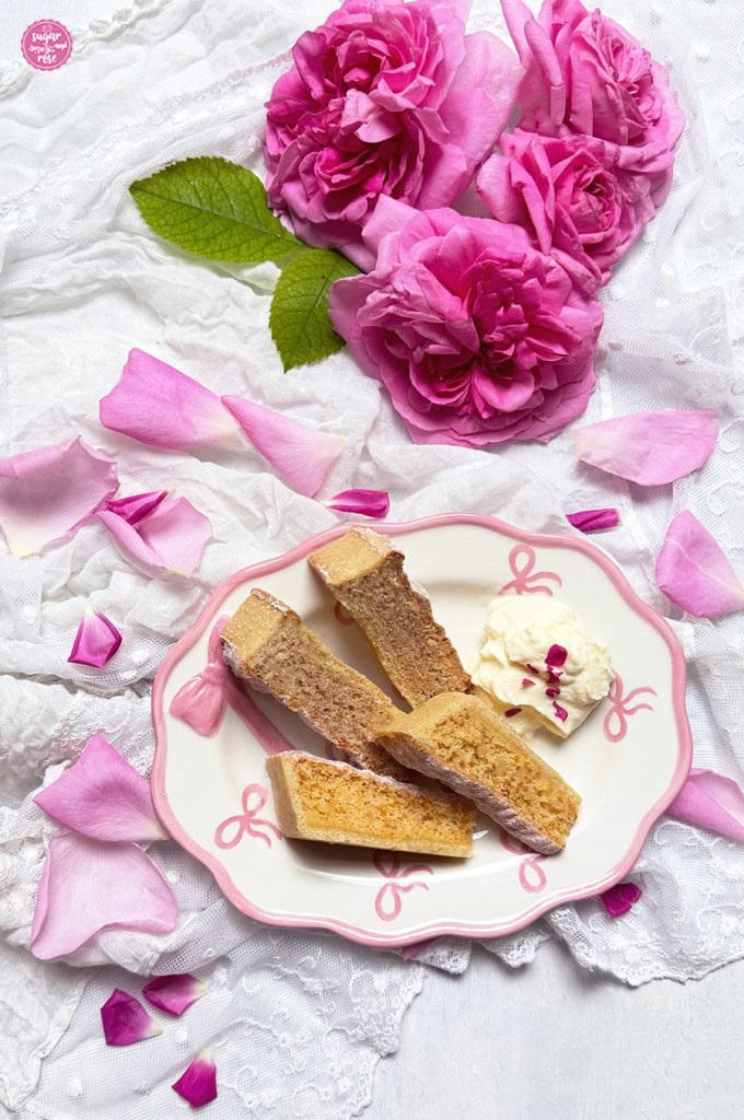 Je zwei Stück der Mandeltarte mit Rosenblüten und der Spanischen Mandeltarte auf einem ovalen Teller mit rosa Dekormaschen und etwas Schlagsahne, rundum liegen rosa Rosenpetalen