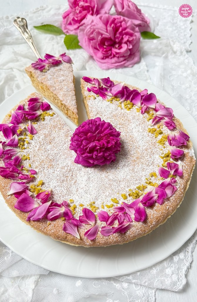Mandeltarte-Variante mit kräftig pinken kleinen Rosenblüten und Pistazien bestreut, davon ein Stück herausgeschnitten und auf einem silbernen Tortenheber abgelegt
