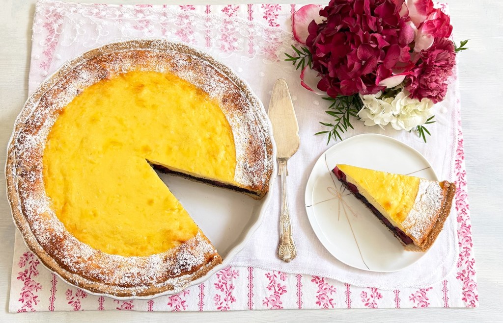 Mohn-Topfenkuchen mit Früchten in einer weißen Keramik-Tarteform, ein Stück ist herausgeschnitten unnd liegt auf einem weißen Porzellanteller mit goldener Masche, gut sichtbar die Mohnschicht und die Kirschen, daneben ein silberner Tortenheber und eine burgunderfarbene Hortensienblüte