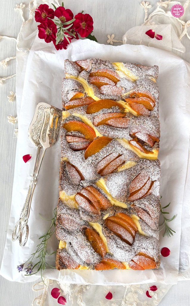 Mohn-Topfenkuchen mit Marillen auf Backpapier, daneben eine silberne Kuchenzange und kleine rote Röschen