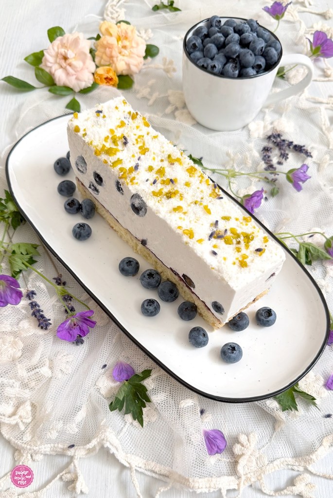 Eine lange Kuchenschnitte auf ovalem Teller, rundum Heidelbeeren, daneben lila Storchenschnabel-Blüten, Lavendelblüten und apricotfarbene Blüten der Rose Garden of Roses